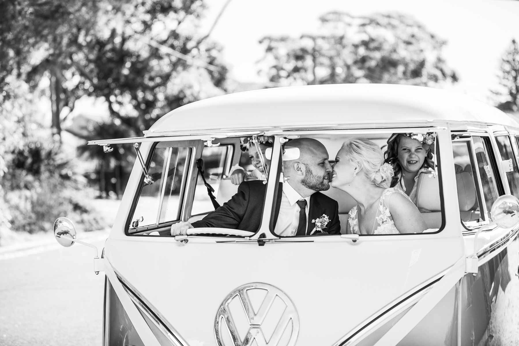 A married couple share a kiss sitting in their wedding Kombi as their kids pull faces