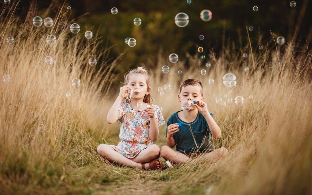 Siblings sit in long grass, blowing bubbles at sunset