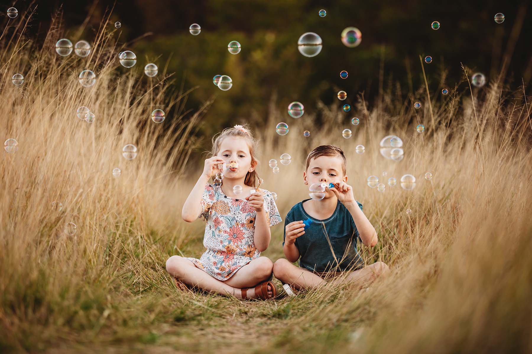 Siblings sit in long grass, blowing bubbles at sunset