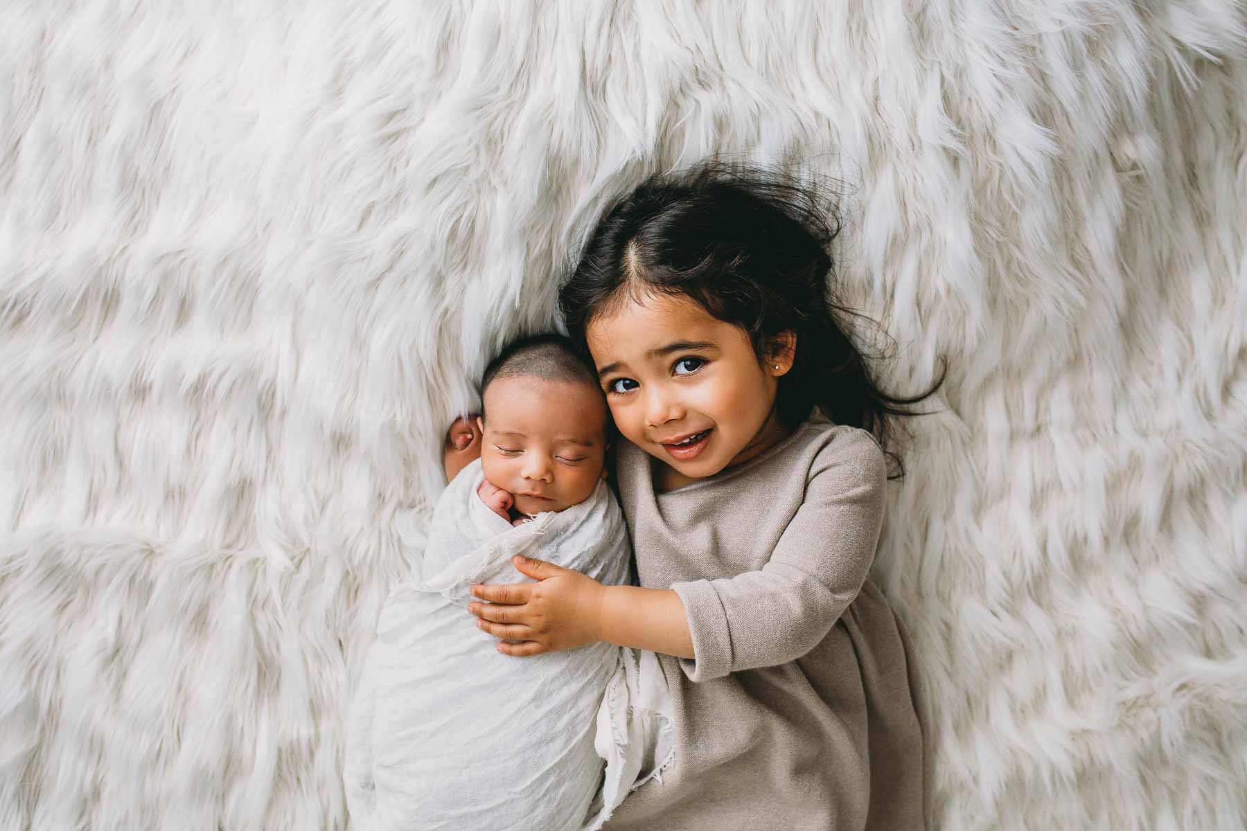 A little girl cradles her newborn baby brother in her arm, laying on a white rug