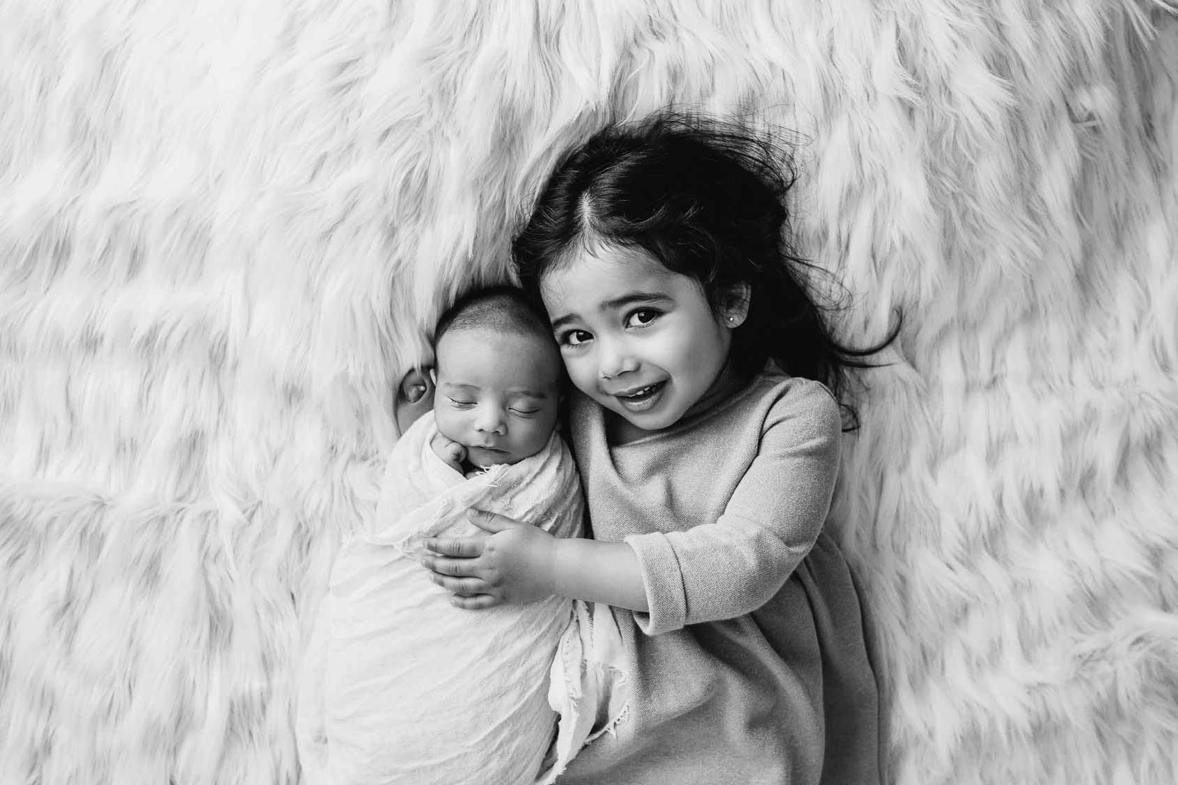 A little girl lays on a rug and cuddles her newborn baby brother