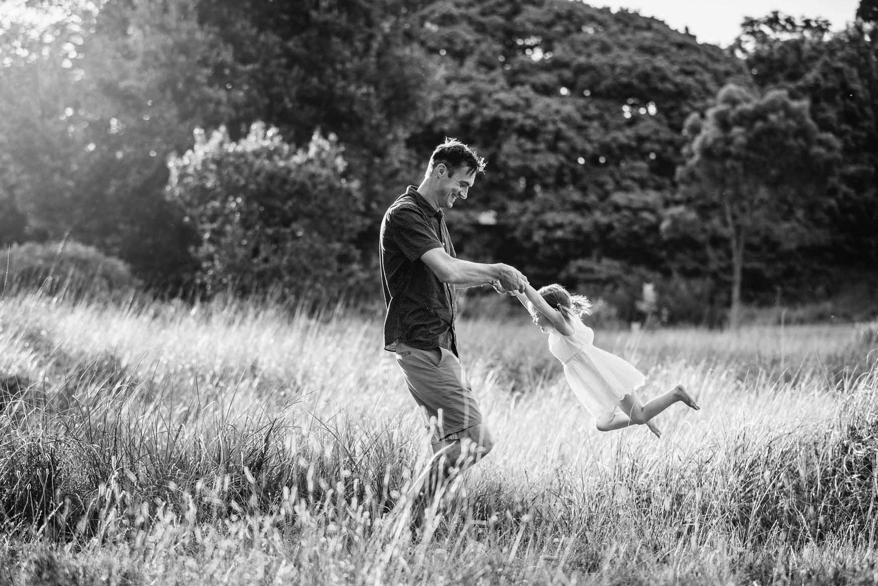 A father holds his daughter's hands and spins her around, in long grass at sunset