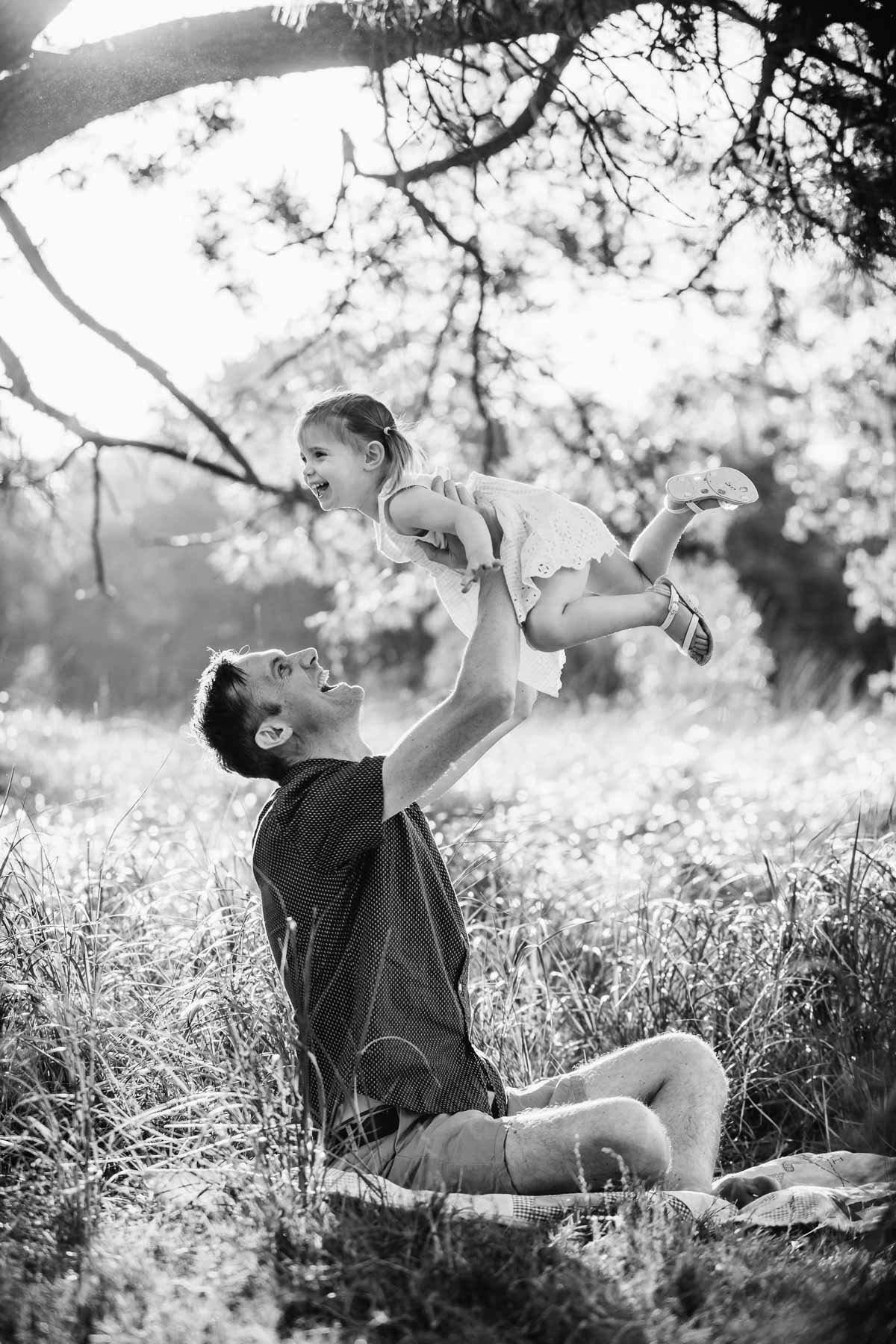 A dad holds his little girl above him as they sit in long grass, sun set behind them