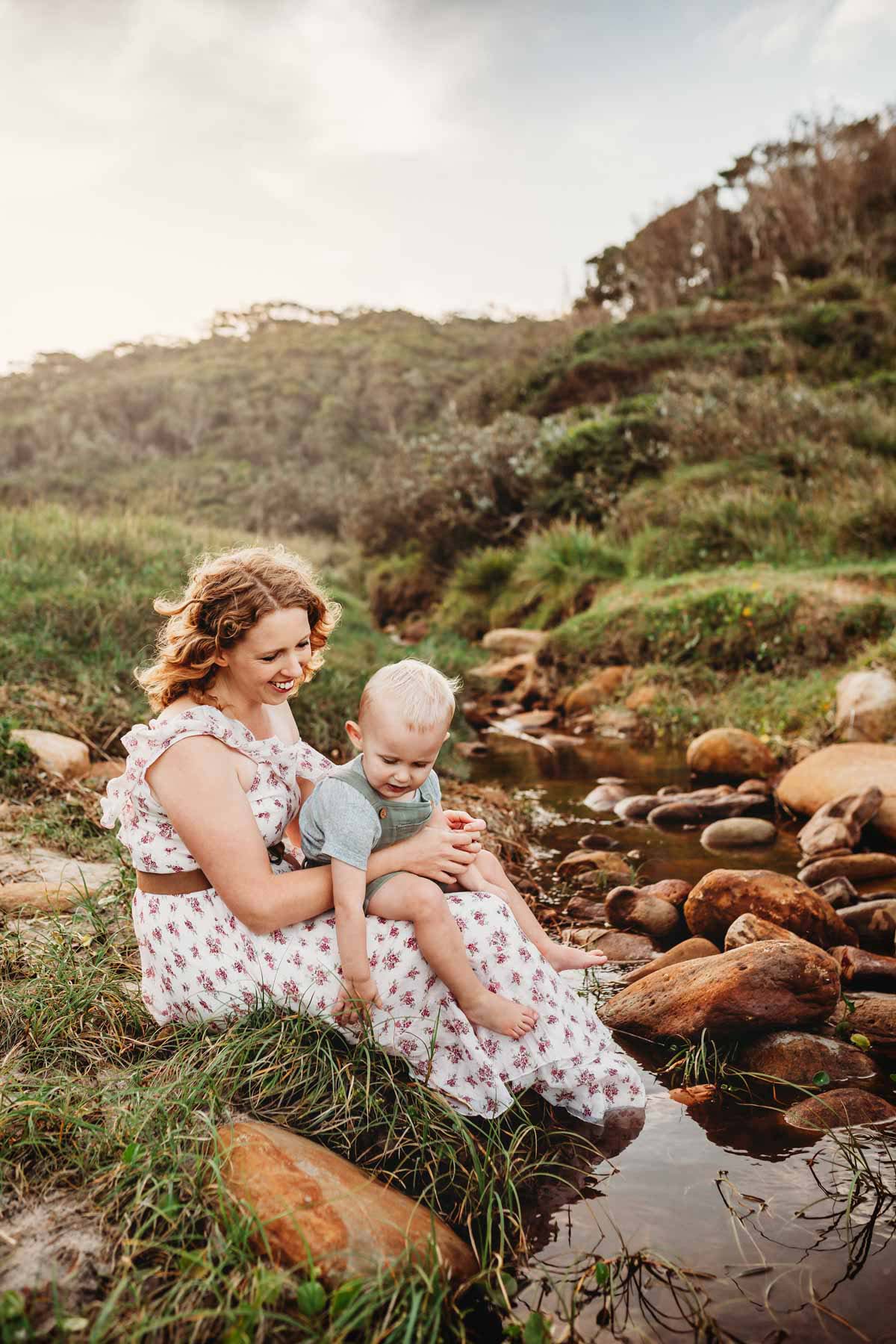 A mother sits on rocks at the beach and holds her baby boy in her lap