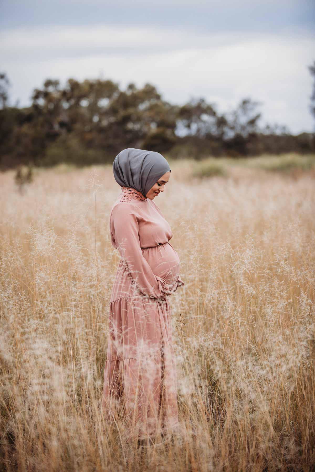 A woman looks down at her pregnant belly as she stands in long grass at sunset