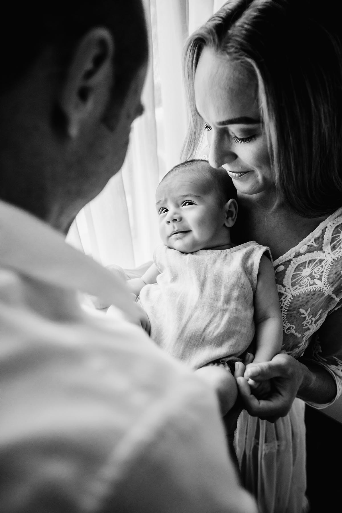 Parents stand together holding their newborn between them, in front of window light