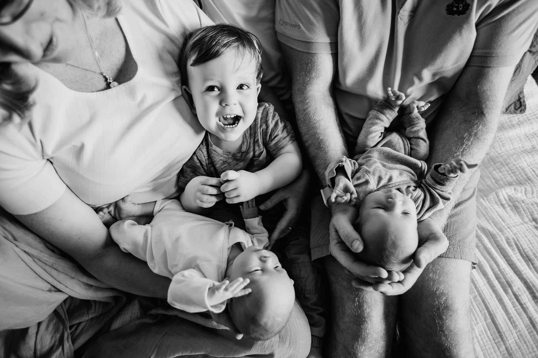 A little boy smiles at the camera as his parents hold their twin newborn babies in their hands