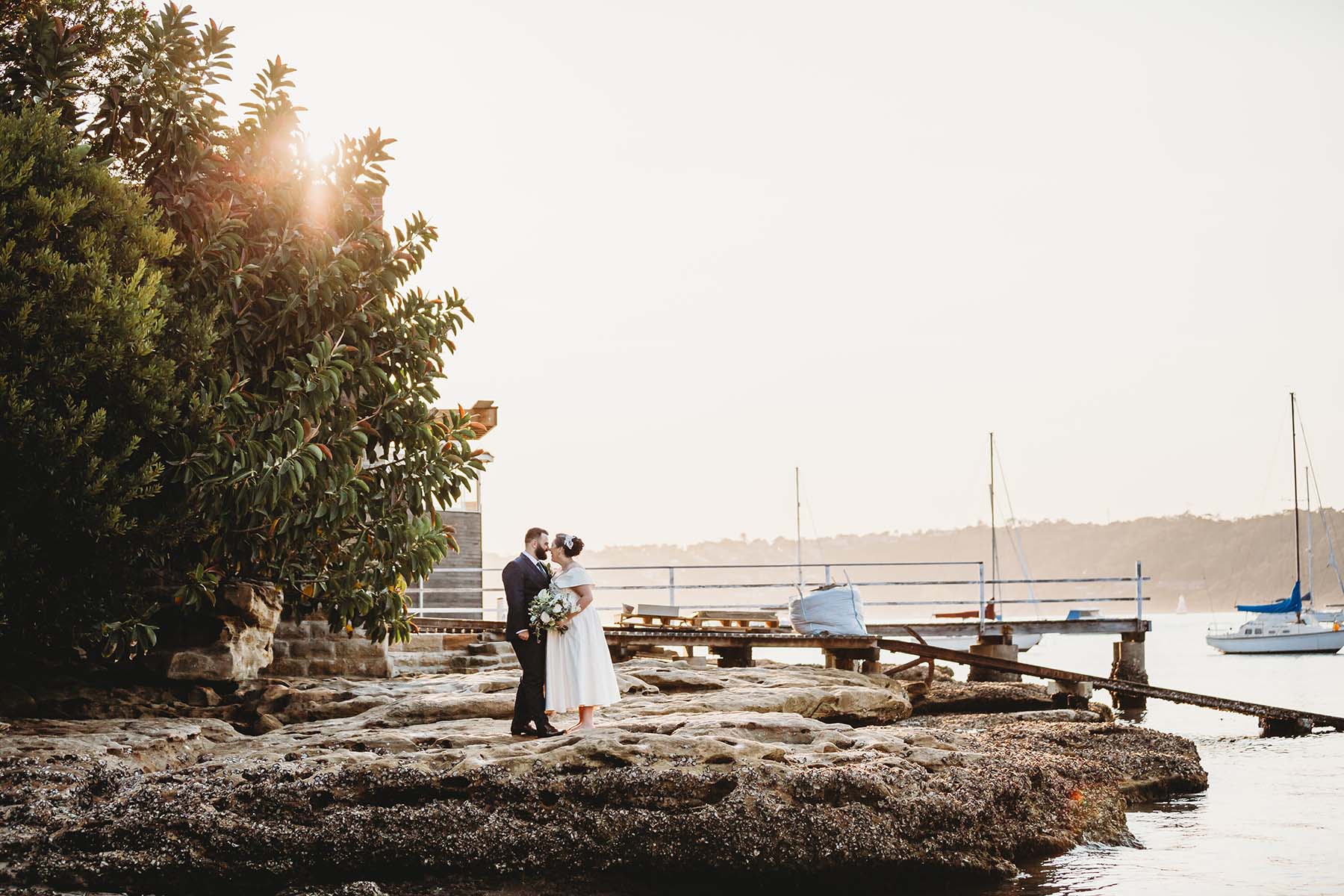 Married couple stand cuddling on beach rocks after their Dunbar House wedding