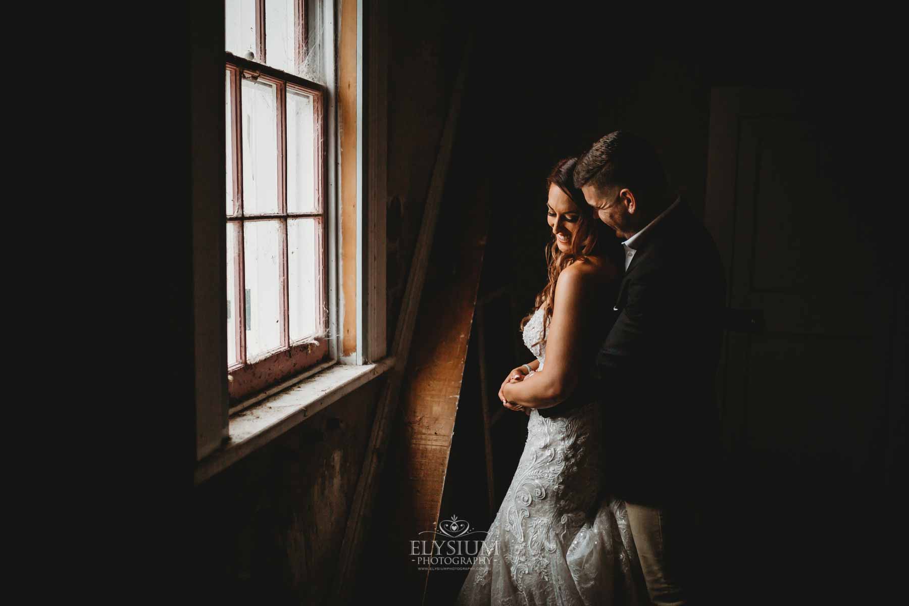 Gledswood Wedding - A groom cuddles his bride in a dimly lit room in front of a small window