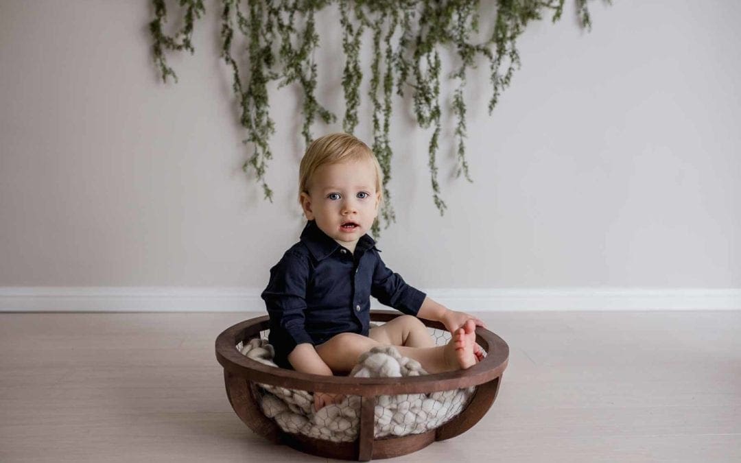 studio image of a baby boy, seated in a wooden bowl, greenery draped behind him