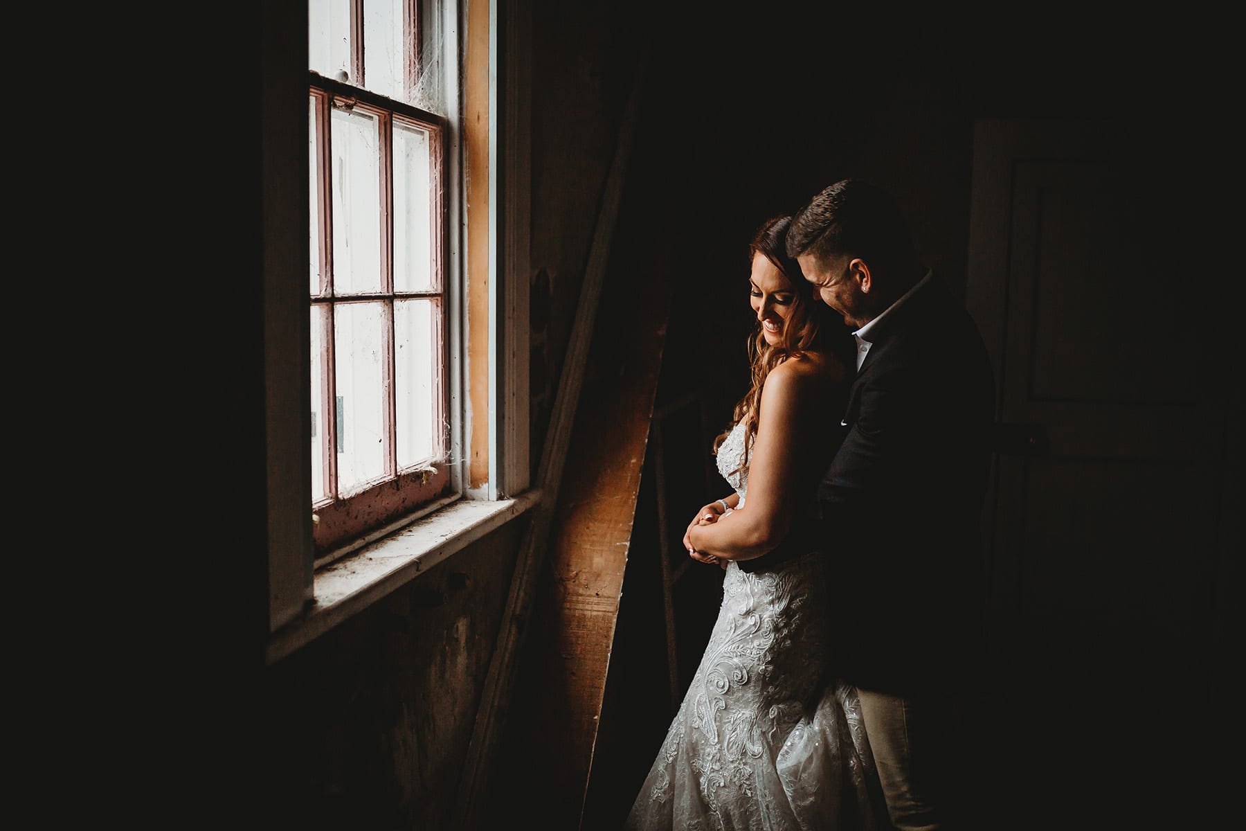 Bride and groom standing in Gledswood Homestead, lit by a small window as they cuddle
