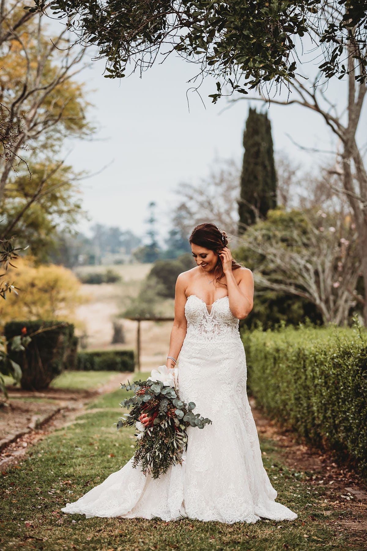 Elysium-Photography-Gledswood-Wedding-Photographer-Gledswood-Estate-T&T-170 A bride brushes her hair behind her ear, standing in a green garden at Gledswood