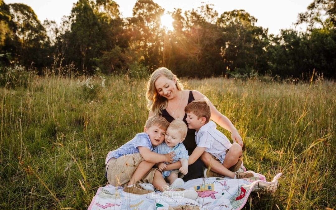 A family sit on a blanket in a grassy field, mother cuddles her 3 boys