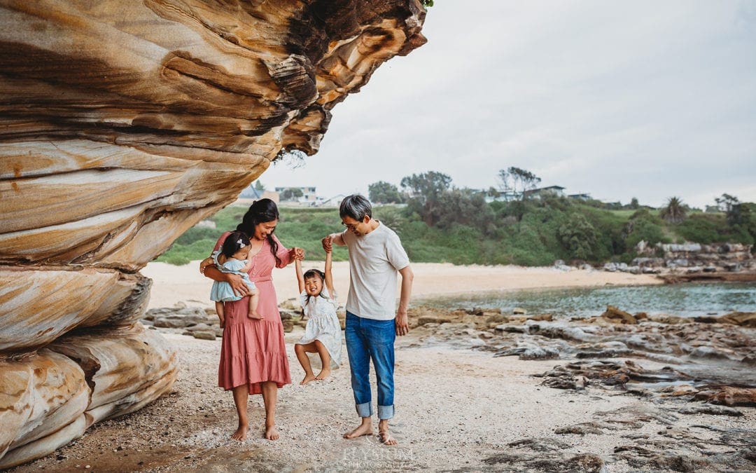 Family Photographer - parents swing their little girl between them as they stand on the beach