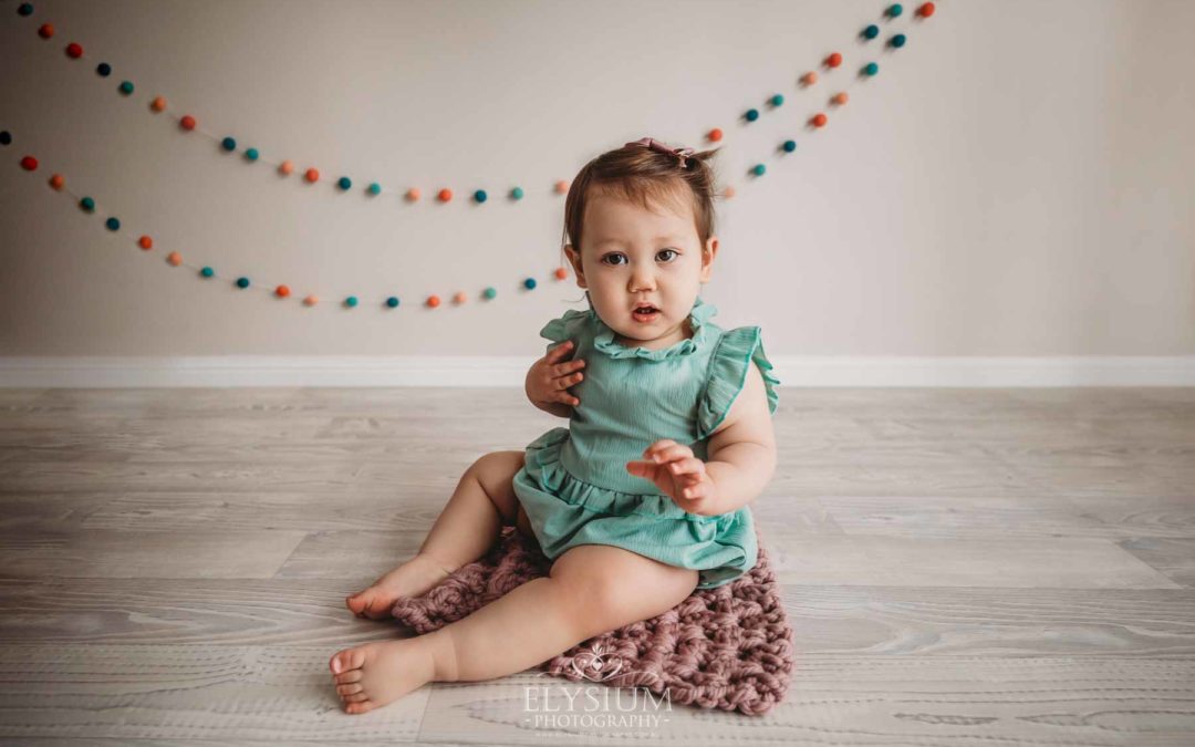 Baby girl wearing a green romper sitting on a pink rug with coloured bunting behind her