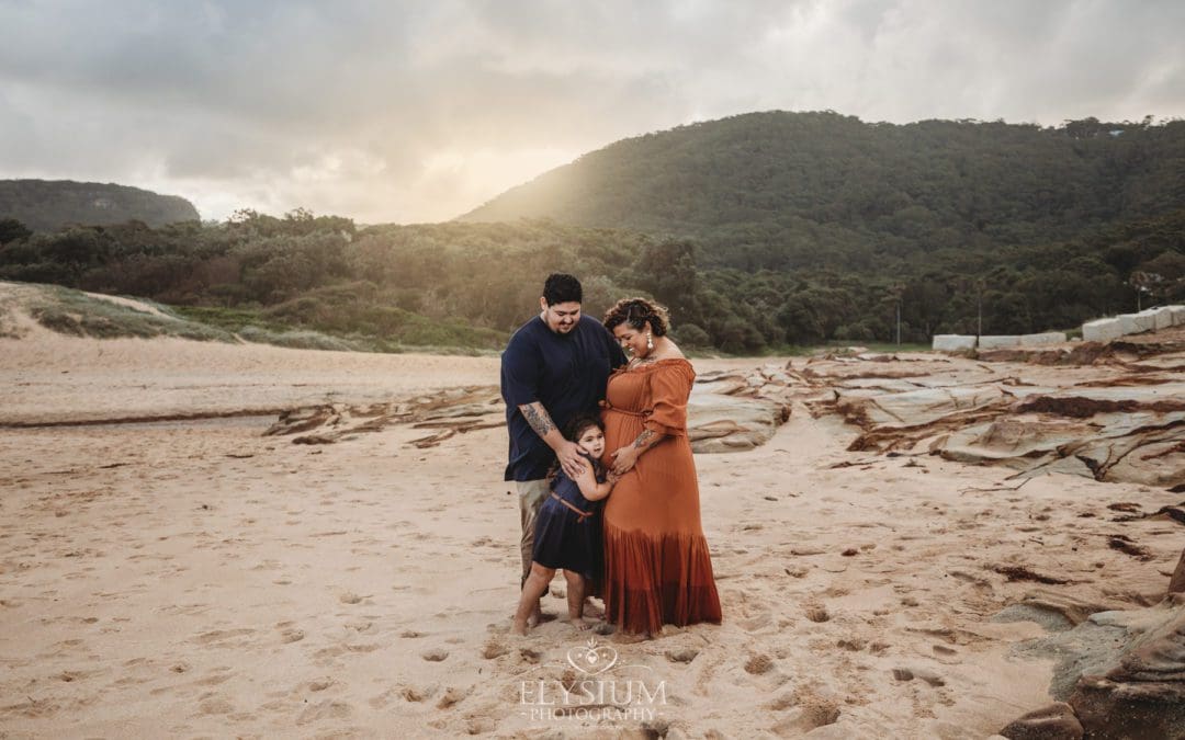 Parents cuddle their baby girl as they stand on a beach with the sun setting behind them