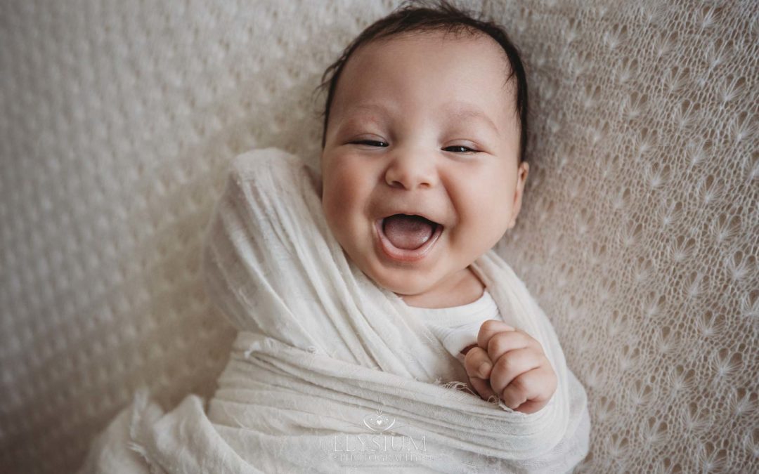 A smiley newborn baby boy lays on a white textured blanket in a soft white wrap