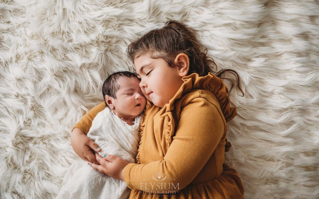 A little girl cuddles her baby sister as they lay on a fluffy white blanket