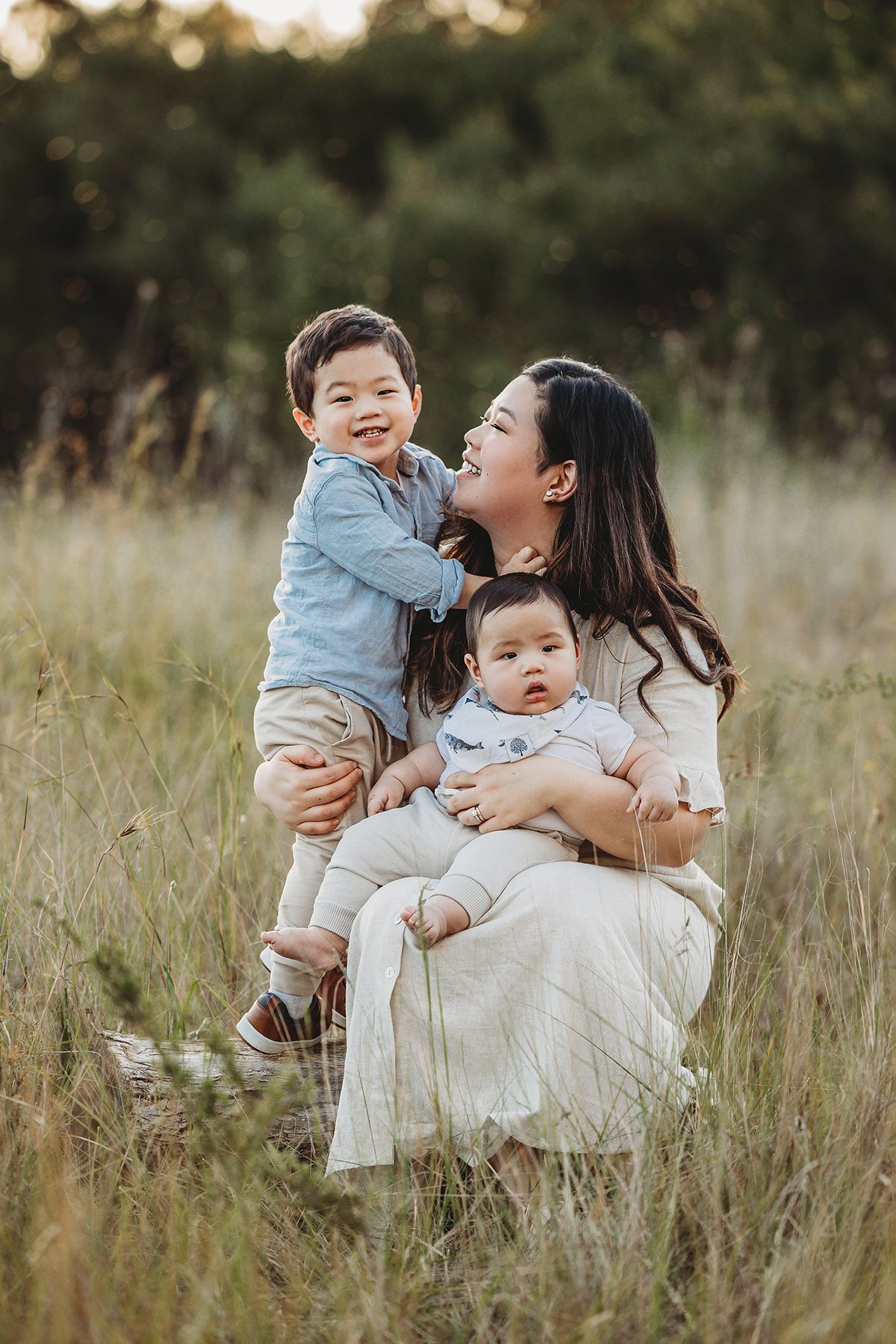A mother sits in a grassy field with her 2 little boys sitting in her lap
