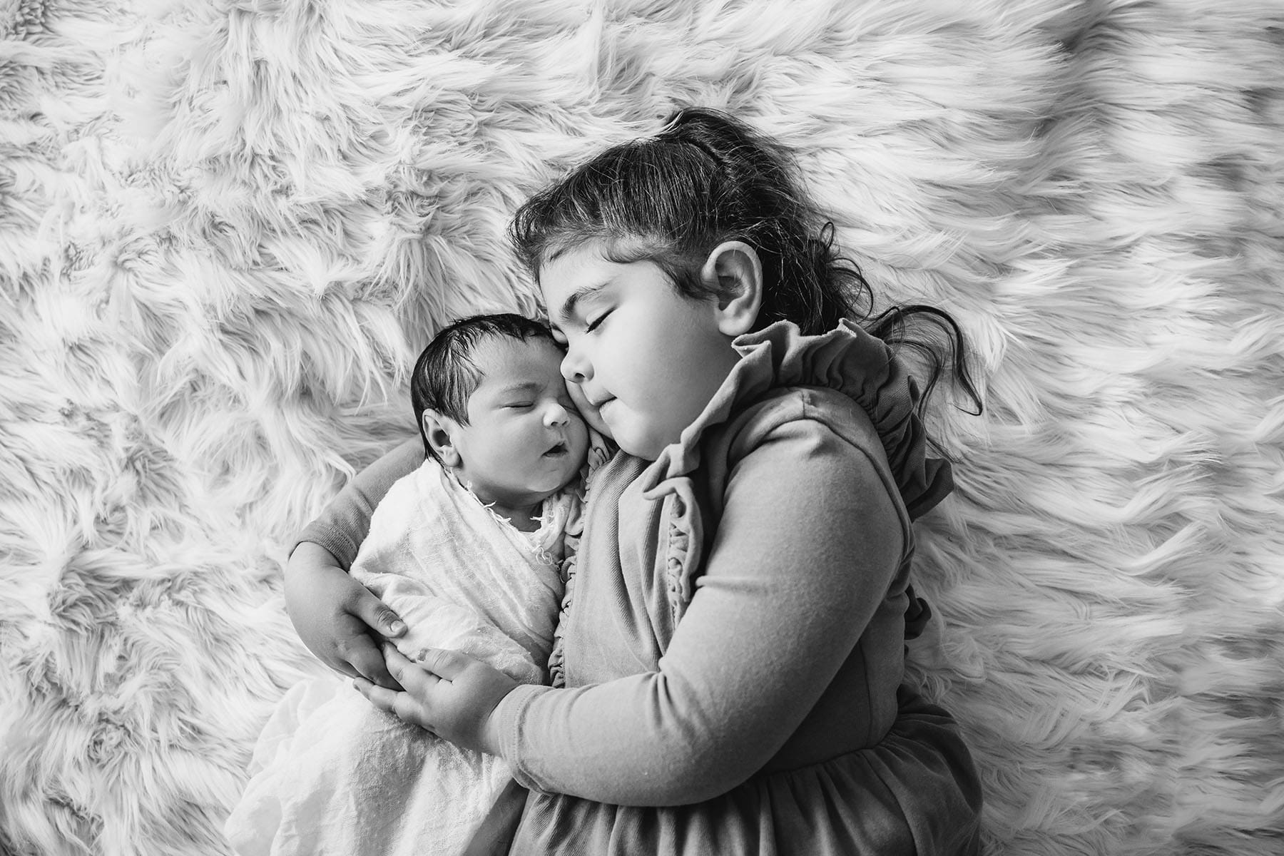 A little girl and a newborn baby lay snuggled together on a white rug