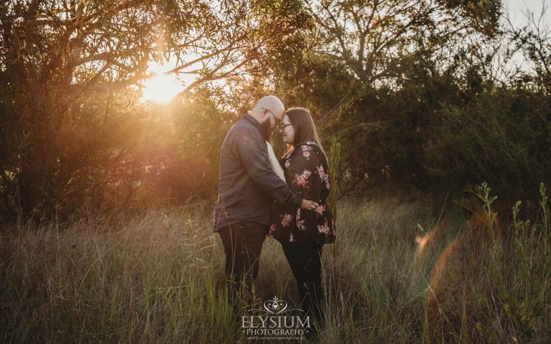 An engaged couple stand hugging in a long grassy field at sunset