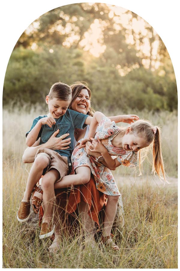 A mother tickles her children as they sit in her lap surrounded by a grassy field at sunset