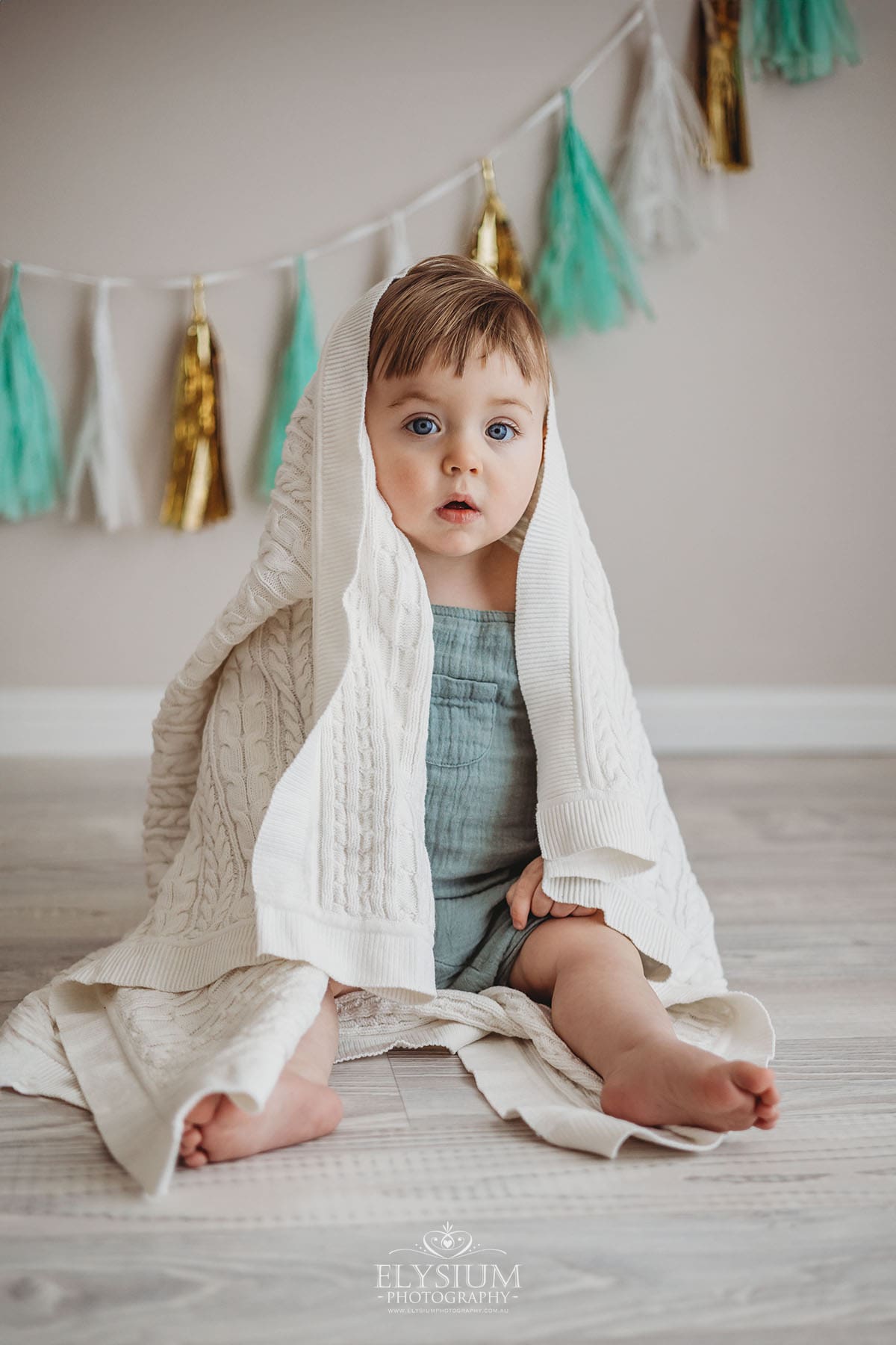 A baby boy sits under a white knitted blanket looking out with big blue eyes in a bright studio