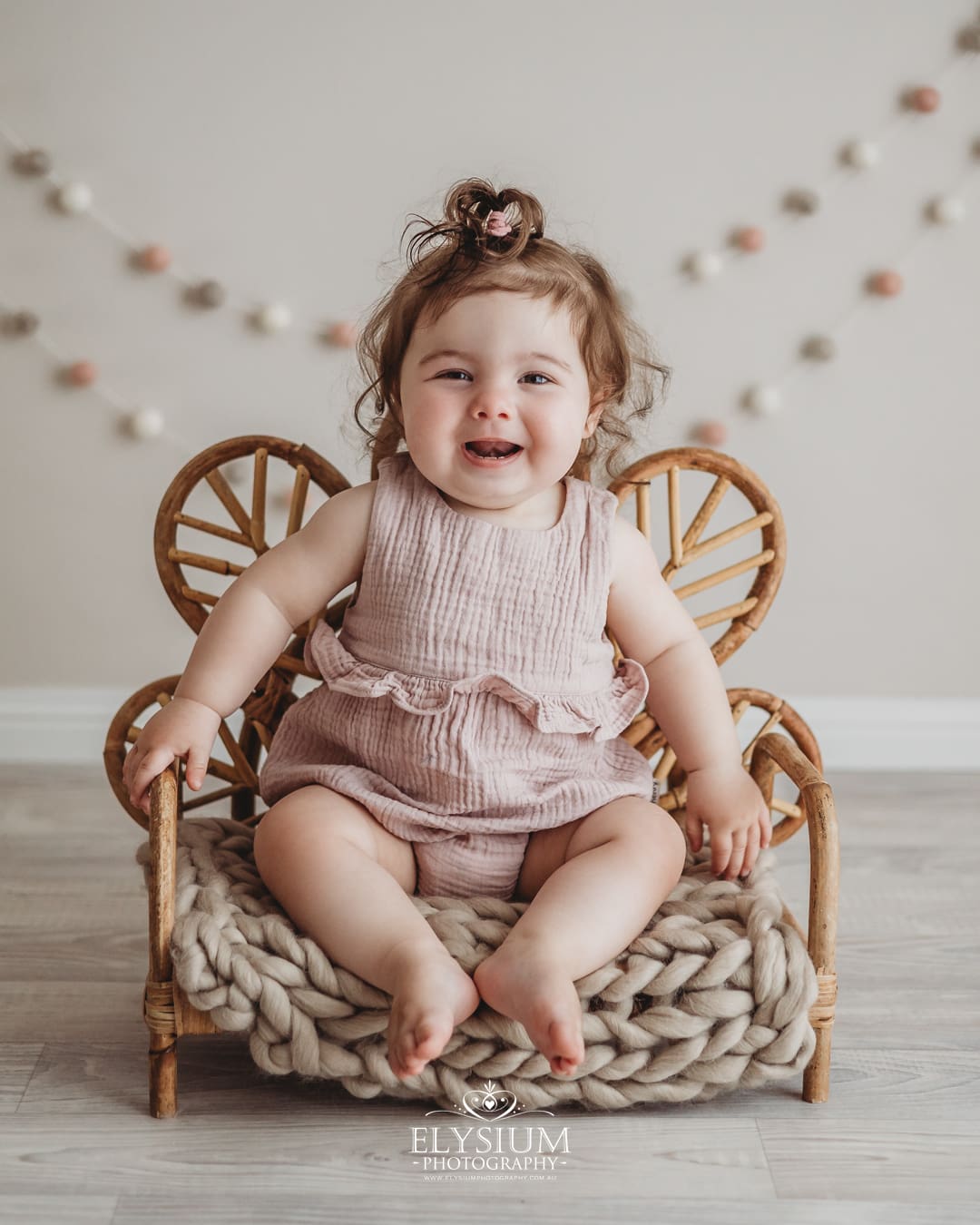 A baby girl sits on a bamboo chair prop wearing a pink romper in a bright studio