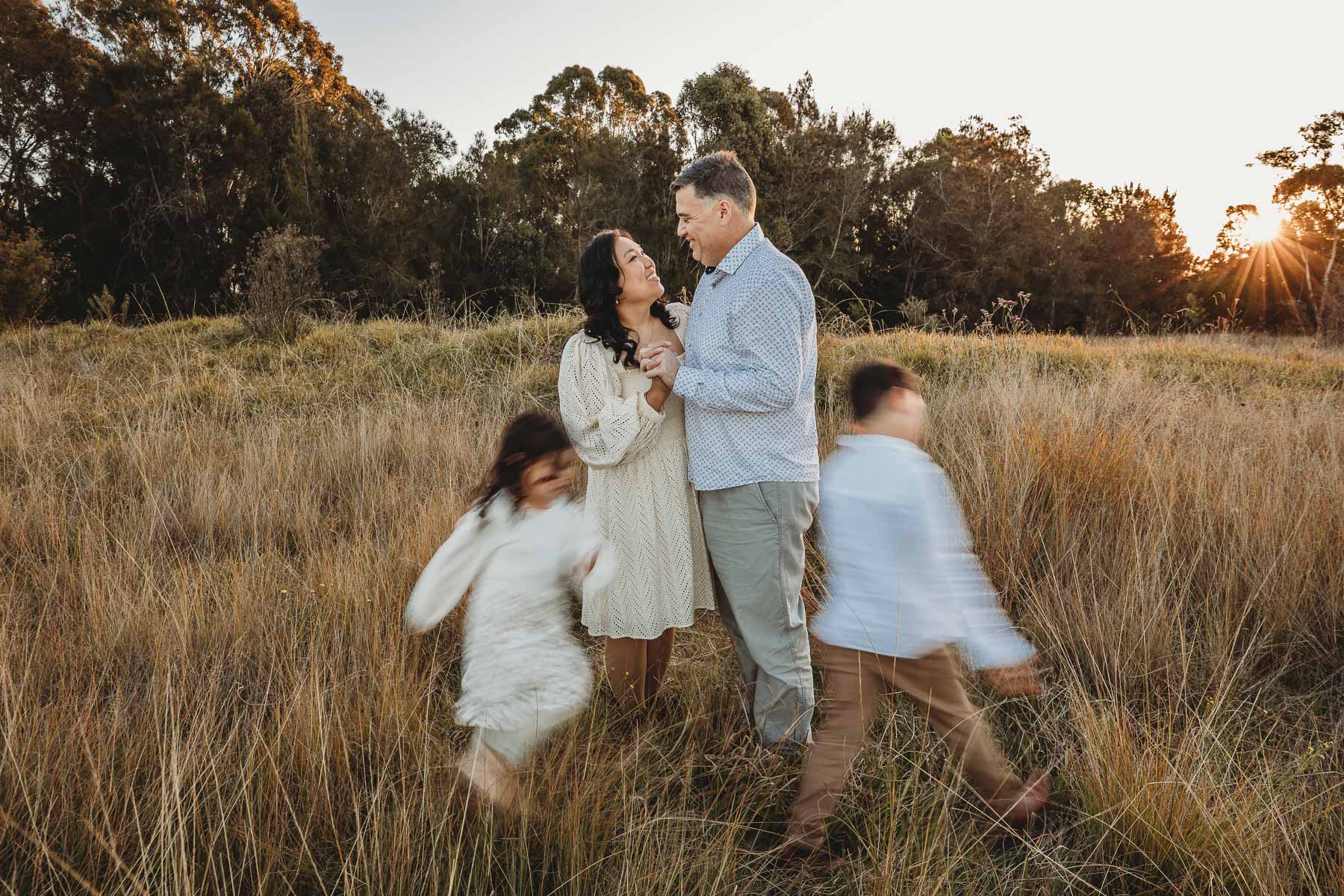 Parents stand in a grassy field as their kids run around them at sunset during their family photo session