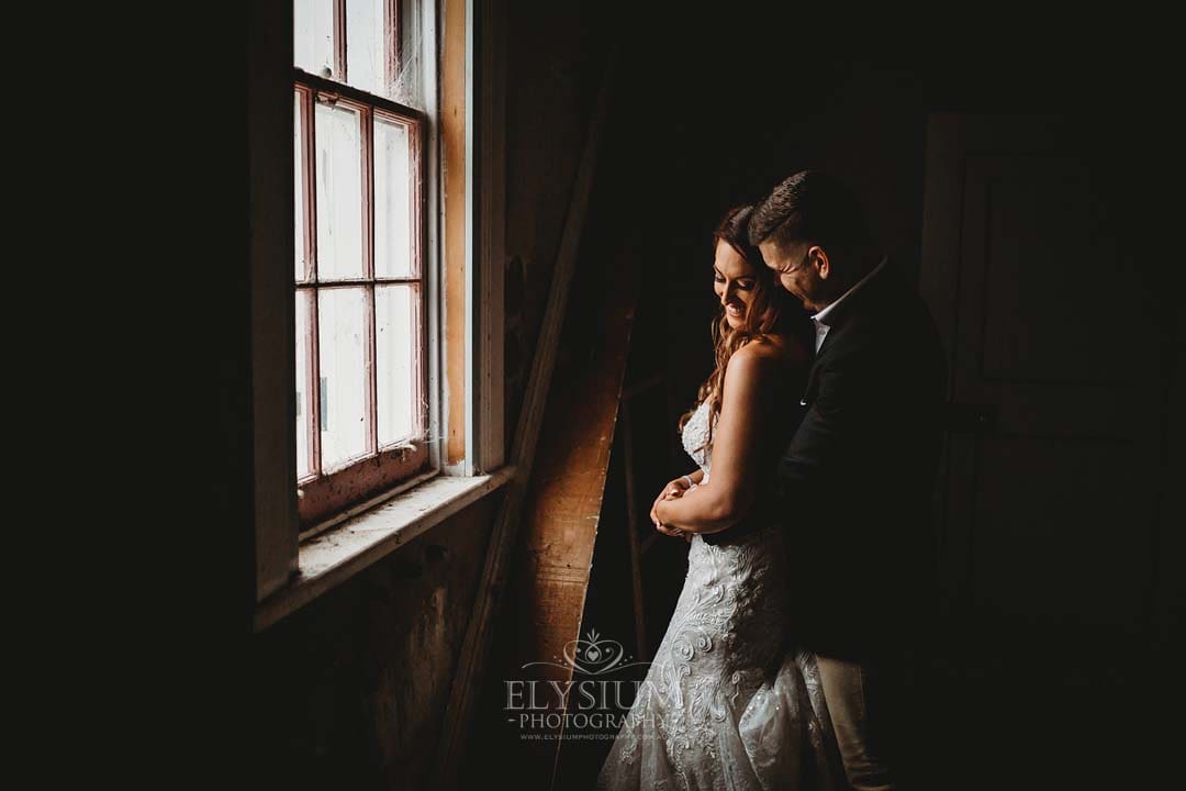 Elysium-Photography-Gledswood-Wedding-Photographer-Gledswood-Estate-T&T-156 A couple smile as they cuddle next to an old window in a darkened room of a homestead