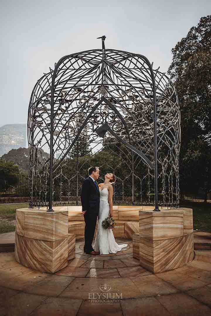 Elysium-Photography-Sydney-Wedding-Photographer-EdLaura-29 A married couple stand hugging under an iron rotunda as rain begins to fall on them