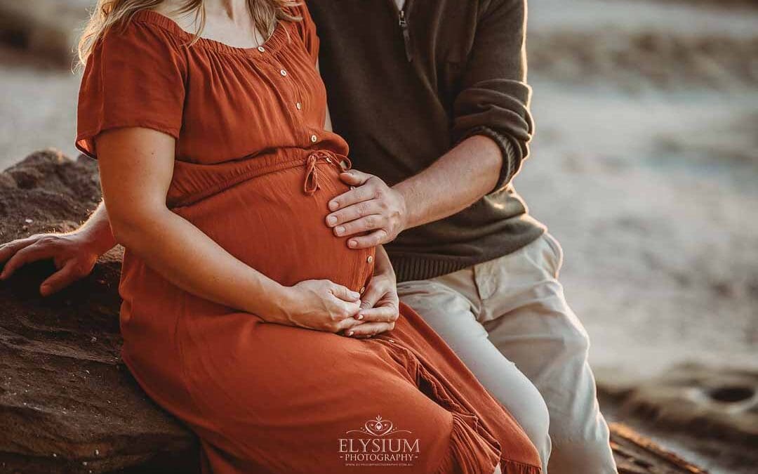 A husband holds his wife's pregnant belly as they sit on a beach rock at sunset
