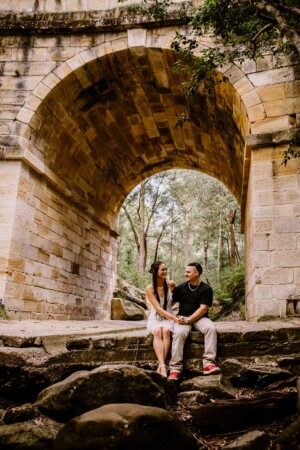 An engaged couple sit under Lennox Bridge holding hands