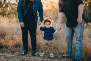 A little boy stands holding his parents hands as the sun sets behind them