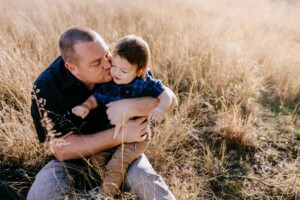 A father kisses his little boy as they sit in long grass at sunset