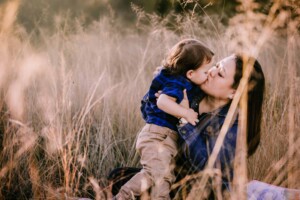 A mother kisses her little boy as they sit in long grass at sunset