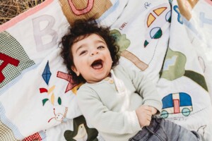 A baby boy giggles and smiles at the camera as he lays back on a rug