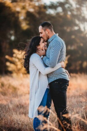 A husband holds his wife's face as he kisses her forehead, standing in a grassy field at sunset