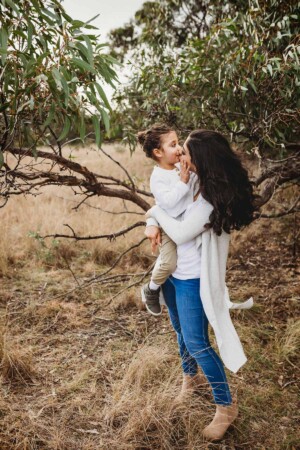 A mother kisses her little boy under a tree in a grassy field