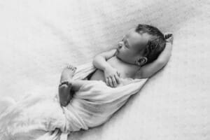 A newborn baby lays sleeping in a white wrap on a textured blanket with his arm above him