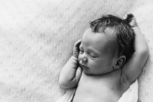 A newborn lays on a patterned white blanket with his arms above him in a white wrap