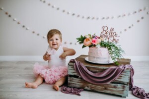 A baby girl dressed in pink sits on a white studio floor next to a cake she is about to smash