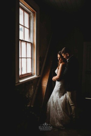 Gledswood Wedding - A groom cuddles his bride in a dimly lit room in front of a small window