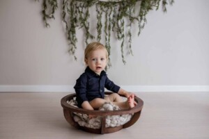 studio image of a baby boy, seated in a wooden bowl, greenery draped behind him
