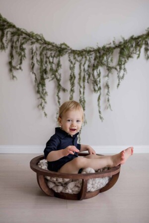 Baby boy smiles as he sits in a wooden bowl in a studio, a hanging green vine behind him