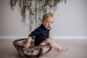 A baby boy sits in a wooden bowl, greenery hanging behind him