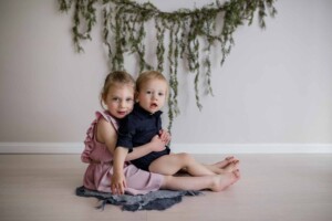 A little girl cuddles her baby brother in her lap, sitting on the floor, green garland hanging behind them