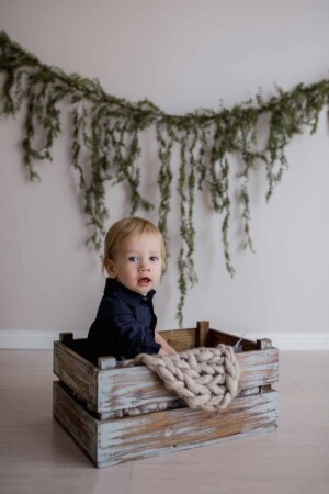 studio image of a baby boy, seated in a wooden crate, greenery draped behind him