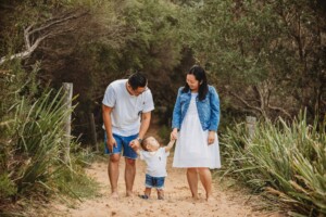 Parents stand on a sandy beach path holding their baby's hands