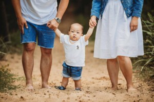 Parents stand on a sandy beach path holding their baby's hands